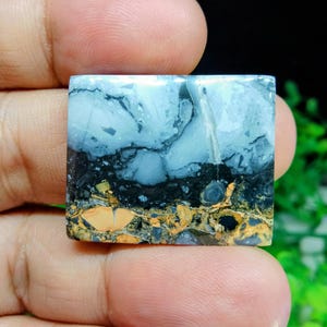 Puede incluir: Una piedra preciosa cuadrada con una apariencia en capas. La capa superior es azul claro con vetas de color gris oscuro, seguida de una capa negra y una capa inferior de patrones moteados de color marr&oacute;n y marr&oacute;n. La piedra se sostiene en una mano.
