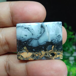 Puede incluir: Una piedra preciosa de forma cuadrada con un patr&oacute;n en capas. La capa superior es azul claro con vetas negras, seguida de una capa negra y una capa inferior de tonos marrones y beige. La piedra se sostiene entre los dedos.