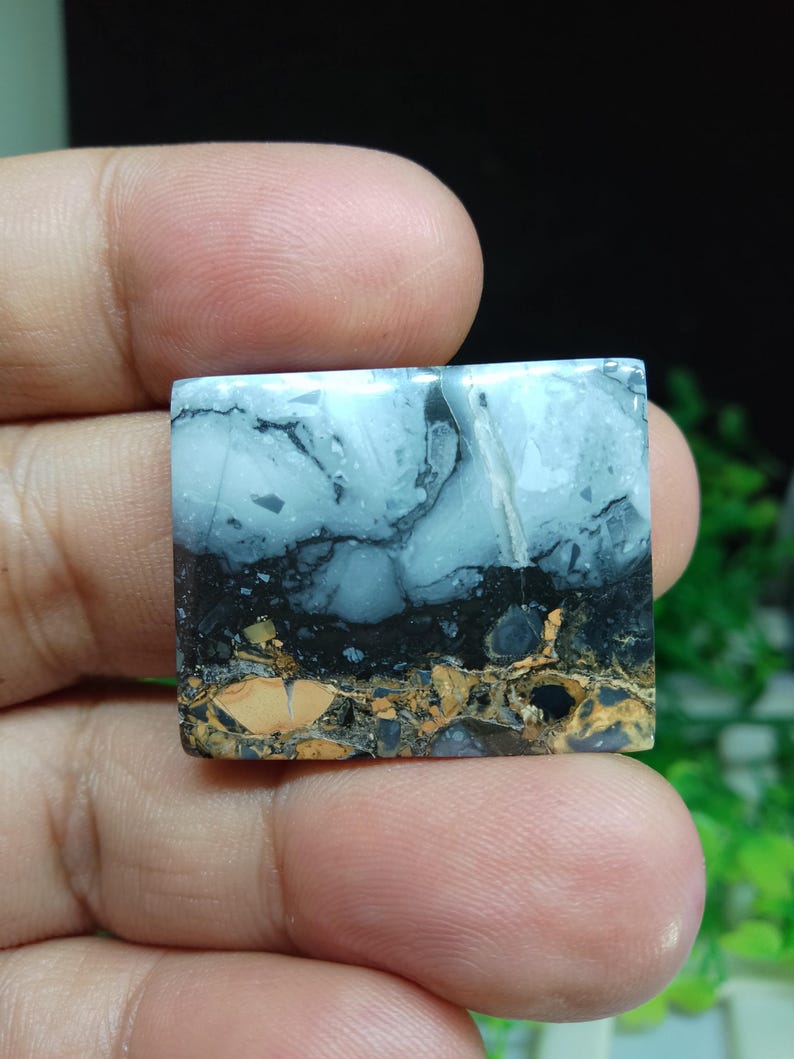 Puede incluir: Una piedra preciosa de forma cuadrada con un patr&oacute;n en capas. La capa superior es azul claro con vetas negras, seguida de una capa negra y una capa inferior de tonos marrones y beige. La piedra se sostiene entre los dedos.
