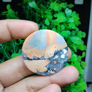 Puede incluir: Una piedra redonda, plana y pulida con un patrón único de marrón, naranja, gris y negro. La piedra tiene un aspecto natural y orgánico.