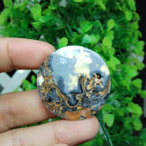 Puede incluir: Una piedra de ágata redonda, plana, gris, marrón y blanca con un patrón de remolino. La piedra tiene un diámetro de aproximadamente 5 cm.