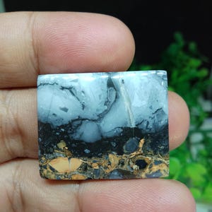 Puede incluir: Una piedra preciosa cuadrada con un aspecto en capas. La capa superior es azul claro con vetas oscuras, seguida de una banda negra y una capa inferior de patrones moteados marrones y beige. La piedra se sostiene en una mano.