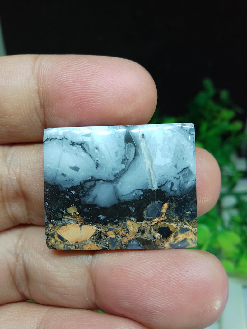 Puede incluir: Una piedra preciosa cuadrada con un aspecto en capas. La capa superior es azul claro con vetas oscuras, seguida de una banda negra y una capa inferior de patrones moteados marrones y beige. La piedra se sostiene en una mano.