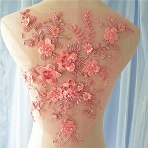 May include: A pink floral applique with 3D flowers, beads, and sequins. The applique features various sizes of flowers and leaves, creating a textured and dimensional design. The applique is likely for embellishing clothing or accessories.