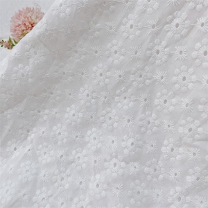 May include: Close-up of white eyelet fabric with a floral pattern. The fabric is soft and textured, with small, embroidered flower designs evenly spaced across the surface. The eyelet fabric is ideal for dresses, blouses, and other apparel.