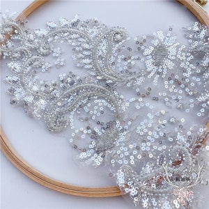 May include: Close-up of a silver lace fabric with intricate beaded and sequined floral embroidery. The delicate design features swirling patterns and sparkling embellishments, perfect for bridal or formal wear. The lace is set against a white background.