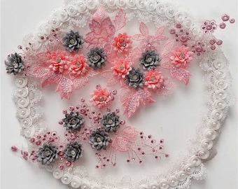 May include: A white lace trim with pink and grey floral embellishments. The trim is decorated with small pearl-like beads.