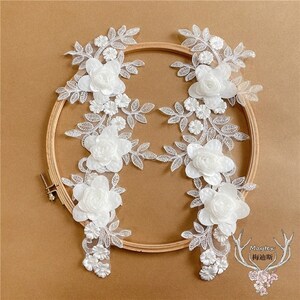 May include: Five white lace appliques with floral designs and 3D flowers. The appliques are arranged in a circle on a wooden embroidery hoop.