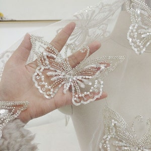 May include: White tulle fabric with delicate silver and pearl butterfly embellishments. The fabric is sheer and has a soft, flowing texture.