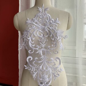 Könnte beinhalten: Weiße Spitzenapplikation mit einem zarten floralen und geschwungenen Design. Die Applikation ist perfekt, um Kleidung, Accessoires oder Heimtextilien einen Hauch von Eleganz zu verleihen.