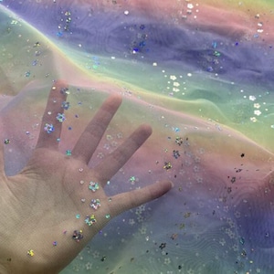 May include: Rainbow tulle fabric with iridescent glitter stars and flowers. The fabric is sheer and has a soft, flowing texture.