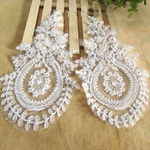 Può includere: Due applicazioni di pizzo bianco con un motivo floreale e un motivo circolare al centro. Le applicazioni sono su uno sfondo di tessuto beige.