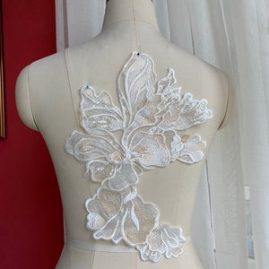 Könnte beinhalten: Weiße florale Spitzenapplikation mit Pailletten. Die Applikation zeigt eine große Blume mit kleineren Blumen und Blättern. Die Applikation ist perfekt, um jedem Kleidungsstück einen Hauch von Eleganz zu verleihen.