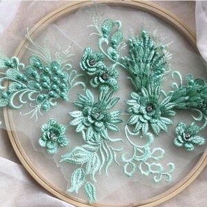 Könnte beinhalten: Nahaufnahme einer zarten grünen Blumenspitze mit Perlen. Die Spitze wird auf einem weißen Hintergrund gezeigt.
