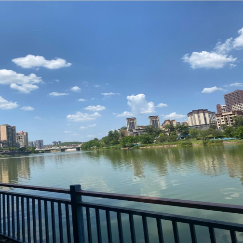 May include: A scenic outdoor view of a lake with buildings in the background under a bright blue sky with fluffy white clouds. A dark blue railing is in the foreground.