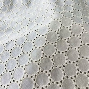 Puede incluir: Tela de algodón blanca con un delicado patrón repetitivo de pequeños círculos. Los círculos están delineados con un hilo blanco y tienen un pequeño agujero en el centro.