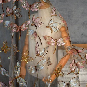 Könnte beinhalten: Transparentes Gewebe mit gestickten Libellen. Die Libellen sind mit schillernden Pailletten in Rosa-, Gold- und Silbertönen verziert. Der Stoff ist über eine Schaufensterpuppe drapiert.