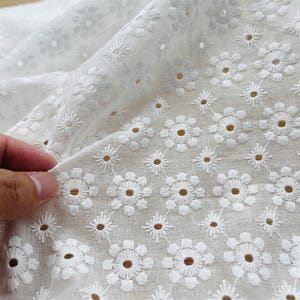 May include: White cotton fabric with a delicate embroidered floral pattern. The fabric has a subtle sheen and a soft, airy feel.