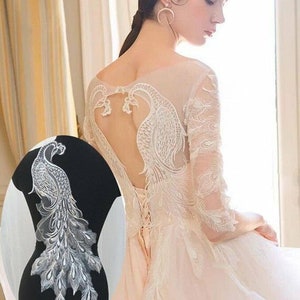 Könnte beinhalten: Ein weißes Spitzenkleid mit einem Pfauenmuster auf dem Rücken. Das Kleid hat einen tiefen Rücken und ein figurbetontes Oberteil. Das Pfauenmuster besteht aus silbernen Pailletten und Perlen.