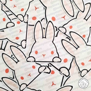 Puede incluir: Primer plano de una pila de pegatinas de conejito blanco con mejillas rosadas y contornos negros. Las pegatinas están dispuestas en un patrón aleatorio y presentan el texto "@bunnydaystore" repetido en todas partes.
