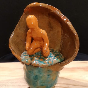 Peut inclure: Une sculpture en céramique d'une personne assise sur une base bleu-vert avec un couvercle brun. La sculpture est en argile et a un aspect rustique et fait main.