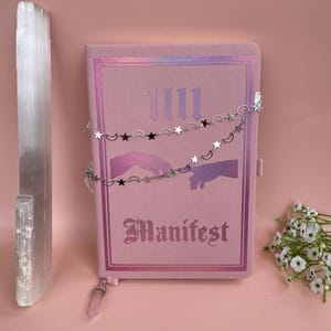 May include: A pink journal with the word "Manifest" printed in a gothic font. The journal has a silver chain with stars and crescent moons hanging from it. The journal is sitting on a pink surface.
