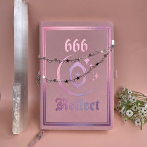 May include: A pink journal with the number 666 and the word "Reflect" in gothic script. A silver chain with stars and moons drapes across the cover. A selenite crystal and a quartz crystal are also present.