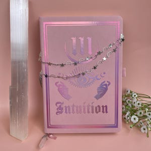 May include: A pink journal with a holographic design featuring the Roman numeral III, a crescent moon, and the word "Intuition". The journal is decorated with a silver chain with stars and crescent moons.