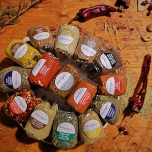 May include: A collection of 15 small, clear plastic bags filled with various spices. The bags are arranged in a circle on a wooden surface. Each bag has a white label with black text, listing the name of the spice. The spices include:  Rosemary, Paprika, Garlic Powder, Chili Flakes, Parsley, Turmeric, Black Pepper, Oregano, Basil, Thyme, Mustard Seeds, Cumin, and Onion Powder.