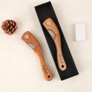 May include: Two wooden bottle openers with a brass accent and a silver blade are shown. One is engraved with "Sophia," the other with "Daniel." They are presented in a black box, with a pinecone and a small white box.