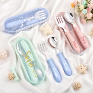 Op de afbeelding: Een verzameling gepersonaliseerde besteksets in pastelkleuren. Elke set bevat een vork en lepel, met namen als "Jason", "Angela", "Sofia" en "William" op de handvatten. De sets zitten in bijpassende etuis, met stervormige decoraties.