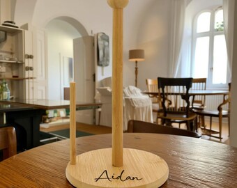 May include: A wooden paper towel holder with a round base and a vertical pole topped with a sphere. The base is engraved with the name "Aidan" in a cursive font. The holder is made of light-colored wood and sits on a wooden table.
