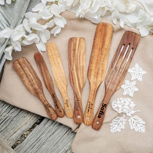 Peut inclure: Ensemble de six ustensiles de cuisine en bois, dont des spatules et une cuillère ajourée, disposés sur une surface beige. Chaque ustensile présente un grain de bois naturel et est gravé d'un nom. Des décorations florales blanches sont en arrière-plan.