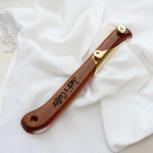 May include: A wooden cutter with brass detailing and the engraved text "Jack's Cutter". The cutter features a curved handle and two brass screws. The cutter is displayed on white fabric.