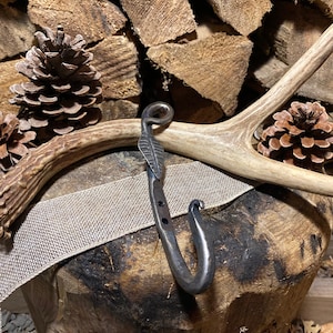 Puede incluir: Un gancho de metal negro con un diseño de hoja, colgado de una asta de ciervo blanca sobre una superficie de madera.