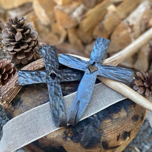 May include: Two black metal crosses with a hammered texture, one with a hole in the centre, are displayed on a wooden surface with a white ribbon.