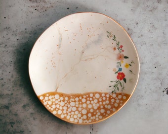 May include: A white ceramic plate with a gold rim and a floral design. The plate has a brown and white speckled pattern around the edge.