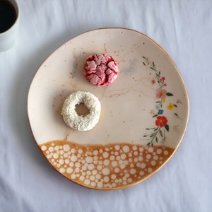 May include: A white ceramic plate with a floral design and a brown speckled pattern around the edge. The plate has two cookies on it, one pink and one white.