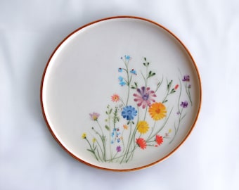May include: A white ceramic plate with a brown rim features a colorful floral design. The flowers are painted in shades of pink, yellow, blue, and purple.