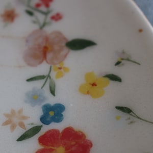 May include: A white ceramic plate with a floral design. The plate features a variety of flowers in shades of pink, yellow, blue, and red. The flowers are painted on the plate and have a delicate, hand-painted look.