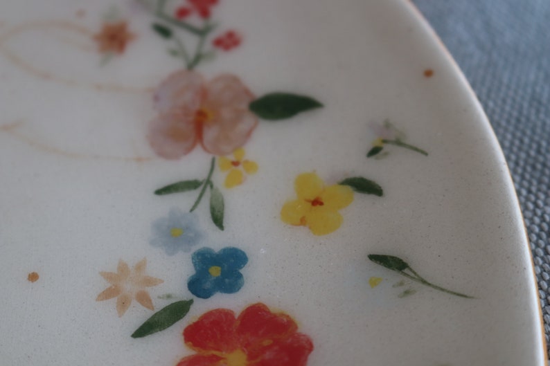 May include: A white ceramic plate with a floral design. The plate features a variety of flowers in shades of pink, yellow, blue, and red. The flowers are painted on the plate and have a delicate, hand-painted look.