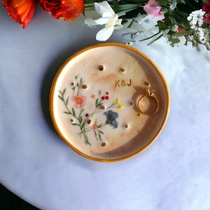 May include: A beige ceramic ring dish with a gold rim, featuring a hand-painted floral design and the initials "K&J." A gold ring rests on the dish. The dish has several small holes.