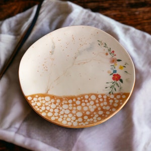 May include: A white ceramic plate with a brown and white speckled design. The plate has a floral pattern with pink, yellow, and red flowers. The plate is on a white fabric background.