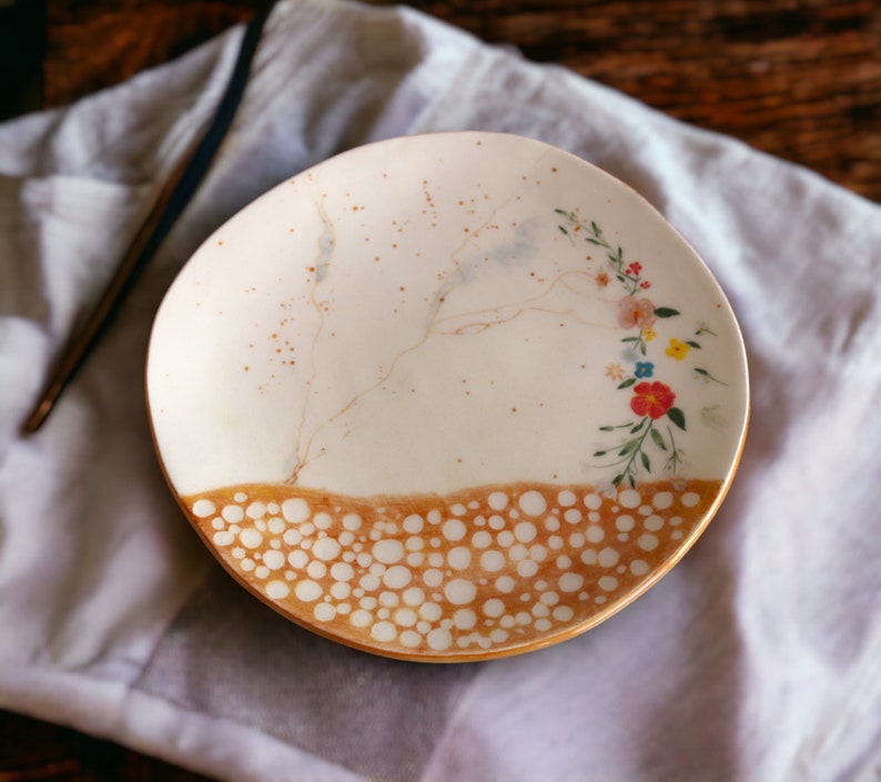 May include: A white ceramic plate with a brown and white speckled design. The plate has a floral pattern with pink, yellow, and red flowers. The plate is on a white fabric background.