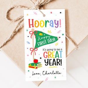 Puede incluir: Una etiqueta de regalo con las palabras "Hooray!" y "Happy First Day!" en letras coloridas. La etiqueta presenta una bandera verde con un lápiz, libros y una manzana. También se incluye el texto "It's going to be a GREAT YEAR!", junto con el nombre "Love, Charlotte."