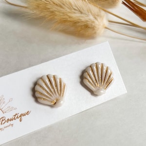 May include: A pair of seashell-shaped stud earrings. The earrings are off-white with gold accents, and are displayed on a white card with the word "Boutique" and the phrase "jewelry" printed in gold. Dried flowers are in the background.
