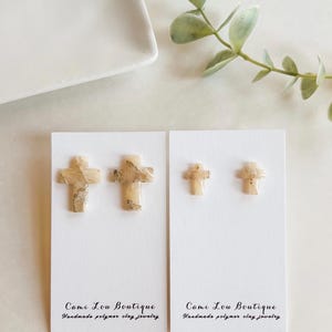 May include: Two pairs of handmade polymer clay cross earrings. One pair is larger than the other. The crosses are beige with brown marbling. The earrings are displayed on white cards with the text "Come Lou Boutique" and "Handmade polymer clay jewelry."
