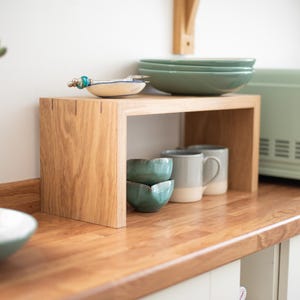 May include: A rectangular light-toned wooden shelf stand with an open design, featuring visible joinery details on the top corners. The stand is positioned on a wooden worktop and holds two small teal bowls and two grey and white ceramic mugs.