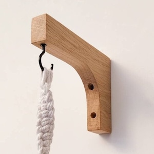 May include: A wooden plant hanger with a natural finish. The bracket has a curved design and a black metal hook. A white macrame plant holder is suspended from the hook. The hanger is mounted on a white wall.