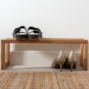 May include: A wooden shoe bench with two pairs of shoes on top. The bench is made of light brown wood and has a simple design. The shoes are a pair of black sandals and a pair of gold high heels.